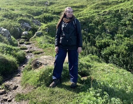 Carla, a young woman, in hiking gear, on a mountainside, surrounded by grass and rocky terrain.