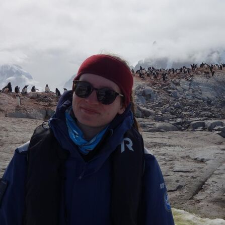 Kate, a young woman, wearing sunglasses, a coat, a lifejacket, and a fleece headband, smiling in front of a penguin colony in Antarctica.